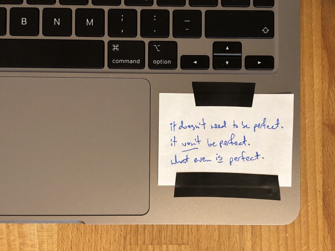 A note stuck to a laptop with the text "It doesn't need to be perfect. It won't be perfect. What even is perfect."